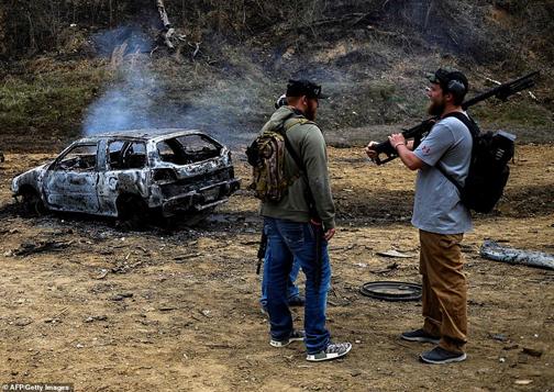 A visitors holding an unloaded gun stands talking to friends near a destroyed car on the main firing line during a break in the shooting. Smoke still billows out from the bonnet of the vehicle and parts of it lay scattered meters away